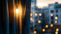 Glowing evening city lights through rain-streaked window with translucent curtain creating dreamy bokeh effect, moody atmospheric urban scene at dusk.