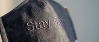Dark denim fabric face mask with embroidered Stay Safe text, close-up shot showing textile texture and stitching detail for health and safety concepts.
