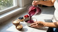 Woman pouring fresh berry smoothie from blender into glass bowl in bright kitchen with healthy ingredients like blueberries and nuts on counter.
