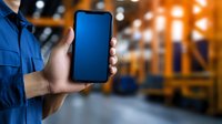 Male warehouse worker in blue uniform holding blank smartphone screen in storage facility with blurred industrial background and bokeh lights.