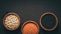 White beans, red lentils, and black lentils in wooden bowls on dark surface. Legumes and pulses for cooking, nutrition, and healthy eating.