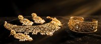 Ornate gold jewelry set with intricate floral patterns including necklace, earrings and bracelet on dark textured surface for luxury fashion accessories.