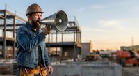 Construction foreman with megaphone directing workers at building site during golden hour sunset with safety helmet and work gear.