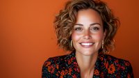 Smiling woman with curly blonde hair wearing floral print blouse against orange background. Professional headshot style portrait showing genuine happiness and confidence.