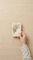 Female hand holding vintage floral greeting card with peach roses and blue flowers on wooden surface, suitable for stationery and greeting card design projects.