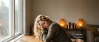 Young blonde woman resting head on arms at wooden table by window in cozy home interior with warm candlelight creating peaceful relaxation moment.