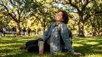 Young woman of mixed ethnicity in casual denim jacket sitting on grass in sunny park with trees, holding colorful beverage bottle, relaxed outdoor lifestyle moment.
