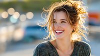 Young Caucasian woman with tousled hair smiling outdoors in golden sunlight, expressing genuine happiness against blurred bokeh background.