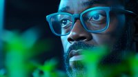 African American man wearing glasses in dramatic blue lighting with green foliage elements in foreground creating artistic portrait composition for business or lifestyle content.