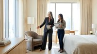 Two professional businesswomen discussing documents in modern hotel room with large windows and neutral decor for corporate travel and meetings.