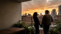 Young couple holding hands on urban rooftop balcony at sunset, overlooking city skyline with golden sky and downtown buildings, romantic evening moment for lifestyle and relationship content.