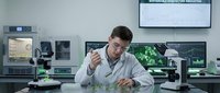 Young male scientist in white lab coat examining plant samples in modern research laboratory with microscopes and computer monitors for biotechnology studies.