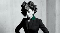 Young woman with geometric black paper origami headdress wearing dark blazer against concrete wall. Modern avant-garde fashion portrait for creative design.
