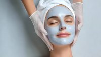Young woman receiving professional facial clay mask treatment at spa salon, beautician hands applying skincare product for rejuvenation.