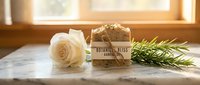 Cream rose with natural soap bar wrapped in kraft paper and fresh rosemary sprig on marble windowsill with warm sunlight, spa and wellness product styling.