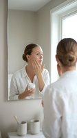 Woman applying moisturizer cream to face while looking in bathroom mirror. Caucasian female in white shirt examining skin, holding cosmetic jar for skincare routine and beauty treatment.