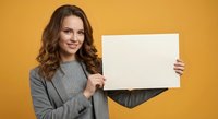 Smiling young Caucasian businesswoman in gray sweater holding blank white paper sign against vibrant yellow background for marketing presentations.