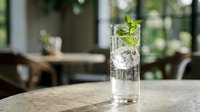 Tall glass of sparkling water with ice cubes and fresh mint sprig on marble table in bright cafe interior.