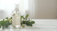 Clear glass spray bottle with natural skincare serum surrounded by fresh eucalyptus leaves and small white flowers on wooden surface for organic beauty products.