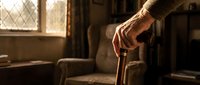 Elderly person's weathered hands gripping wooden walking cane in warm sunlit room, representing aging, support, independence, and senior care concepts.