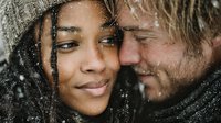 Young mixed-race woman and bearded Caucasian man embracing intimately in falling snow, wearing winter knit hats, romantic couple moment outdoors in cold weather.