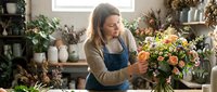 Young female florist arranging colorful bouquet in flower shop workshop with natural lighting and plants in background.