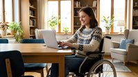 Woman with dark hair and medium complexion using laptop while seated in wheelchair at wooden desk in bright home office with bookshelves, plants, and natural light for remote work and accessibility themes.