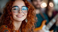 Young caucasian woman with natural red curly hair and glasses smiling genuinely while enjoying music performance, warm bokeh background creates cozy atmosphere.