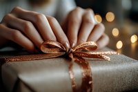 Female hands tying elegant rose gold ribbon on wrapped present against soft bokeh lights background, creating festive holiday atmosphere and magical moment.