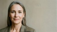Mature woman with long gray hair and fair complexion wearing olive blazer against neutral background, professional headshot for corporate, business, and editorial use.