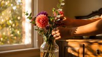 Woman arranging fresh dahlia flowers in glass vase on wooden table by sunny window. Cozy home interior with warm natural lighting for lifestyle content.