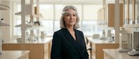 Senior woman with long gray hair wearing black blazer standing in modern bathroom showroom with display fixtures and natural lighting.