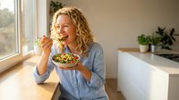 Smiling blonde Caucasian woman enjoying healthy salad bowl in bright modern kitchen. Nutritious eating lifestyle concept for wellness and diet content.