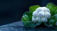 Fresh white cauliflower head with green leaves on dark wet surface with water drops, macro shot showing detailed texture of vegetable in natural lighting.