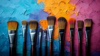 Professional makeup brushes arranged in row against vibrant multicolored textured background with blue, orange and purple paint strokes. Artistic cosmetic tools display.