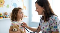 Female pediatrician examining young girl with stethoscope in colorful medical office. Child holds teddy bear during checkup with healthcare professional.