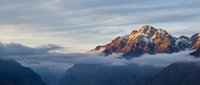 Majestic snow-capped mountain peaks emerging from clouds during golden hour sunrise, dramatic alpine landscape with warm light illuminating rocky summits.