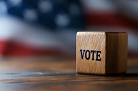 Wooden cube with vote text against blurred American flag background on rustic table, representing democracy, elections and political participation.