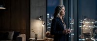Professional businesswoman in elegant black suit holding glass while standing by floor-to-ceiling windows overlooking illuminated city skyline at night.