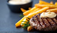 Juicy grilled beef burger patty with creamy sauce and crispy french fries on dark background. Close up view of gourmet fast food meal with rosemary garnish.