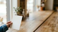 Person with medium skin tone holding blank white card in modern home interior with natural light, suitable for greeting cards, invitations, marketing mockups, and promotional materials.