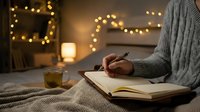 Person writing in journal with warm holiday lights and tea, cozy winter evening atmosphere for wellness and mindfulness content.