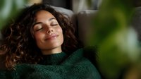 Young Hispanic woman with curly hair relaxing with closed eyes and gentle smile, wearing green knitted sweater against blurred natural background.