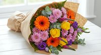 Colorful mixed flower bouquet with orange gerberas, yellow roses, purple chrysanthemums and lisianthus wrapped in kraft paper on white wooden surface.
