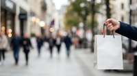Person holding blank white shopping bag on busy urban street with blurred pedestrians and storefronts in background. Retail and commerce concept.