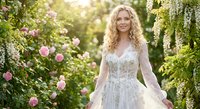 Young blonde woman in elegant white wedding dress standing in blooming garden with pink roses and green foliage during golden hour sunlight.