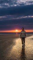 Middle-aged woman with long brown hair walking alone on wet beach sand during dramatic sunset with golden and purple sky, ocean horizon and clouds.