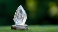 Clear quartz crystal point standing upright on natural stone base against blurred green nature background, showcasing raw mineral specimen in outdoor setting.