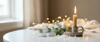 Cozy home interior with burning candle in brass holder, eucalyptus branch and warm fairy lights on white table creating peaceful hygge atmosphere for relaxation.