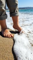 Child's bare feet standing in shallow ocean waves on sandy beach with rolled-up denim pants, enjoying summer vacation and coastal leisure activities.